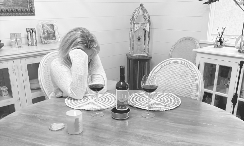 Author sitting at a table by herself with head in her hands. 2 full wine glasses on the table and a bottle of 111 Spanish wine